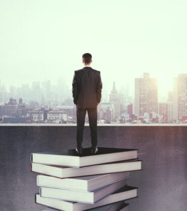 Back view of thoughtful young businessman on rooftop standing on book stack. Bangkok city background. Education and success concept
