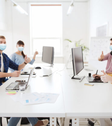 health, safety and pandemic concept - creative team wearing protective medical masks for protection from virus disease with computers showing thumbs up at office
