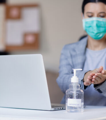 Woman wearing face mask while working in office with laptop and using hand alcohol gel or sanitizer bottle dispenser against corona virus Covid-19. Antiseptic, hygiene and health concept. Prevention of the pandemic virus outbreak