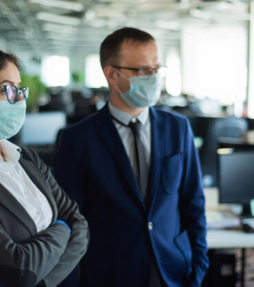 Colleagues in protective masks work in an open space office. A man and a woman in suits work at the workplace. Head and subordinate. European business people during a virus outbreak