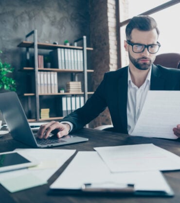 Photo of attentive handsome business brunet guy notebook table reading, paper report before filling online statistics wear black blazer white shirt suit sitting chair office indoors