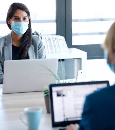 Shot of two business women wearing a hygienic face mask while work with laptops in the coworking space. Social distancing concept.