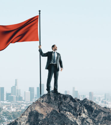 Happy businessman with red flag standing on mountain. City skyline background. Leadership and success concept