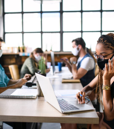 Portrait of young businesspeople with face masks working indoors in office, using laptop and smartphone.