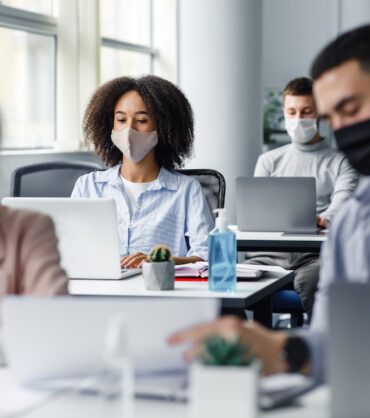 Work with documents and discussion at coronavirus. Guy and woman in protective masks are looking at paper, close up, blurred, focus on african american lady typing on laptop at workplace in office interior