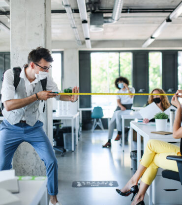 Young people with face masks back at work in office after lockdown, measuring safe distance.