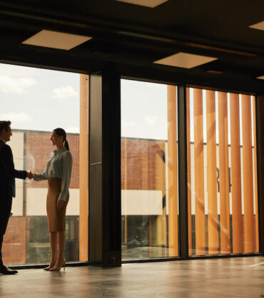 Wide angle view of real estate agent shaking hands with client while standing in empty office building interior lit by sunlight, copy space