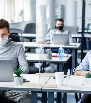 New normal and modern office work during outbreak of coronavirus. Guy and african american woman in protective masks work on workplace with antiseptic and laptops through glass board in interior