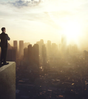Confident businessman standing on the building rooftop while looking at the silhouette of cityscape at dusk time